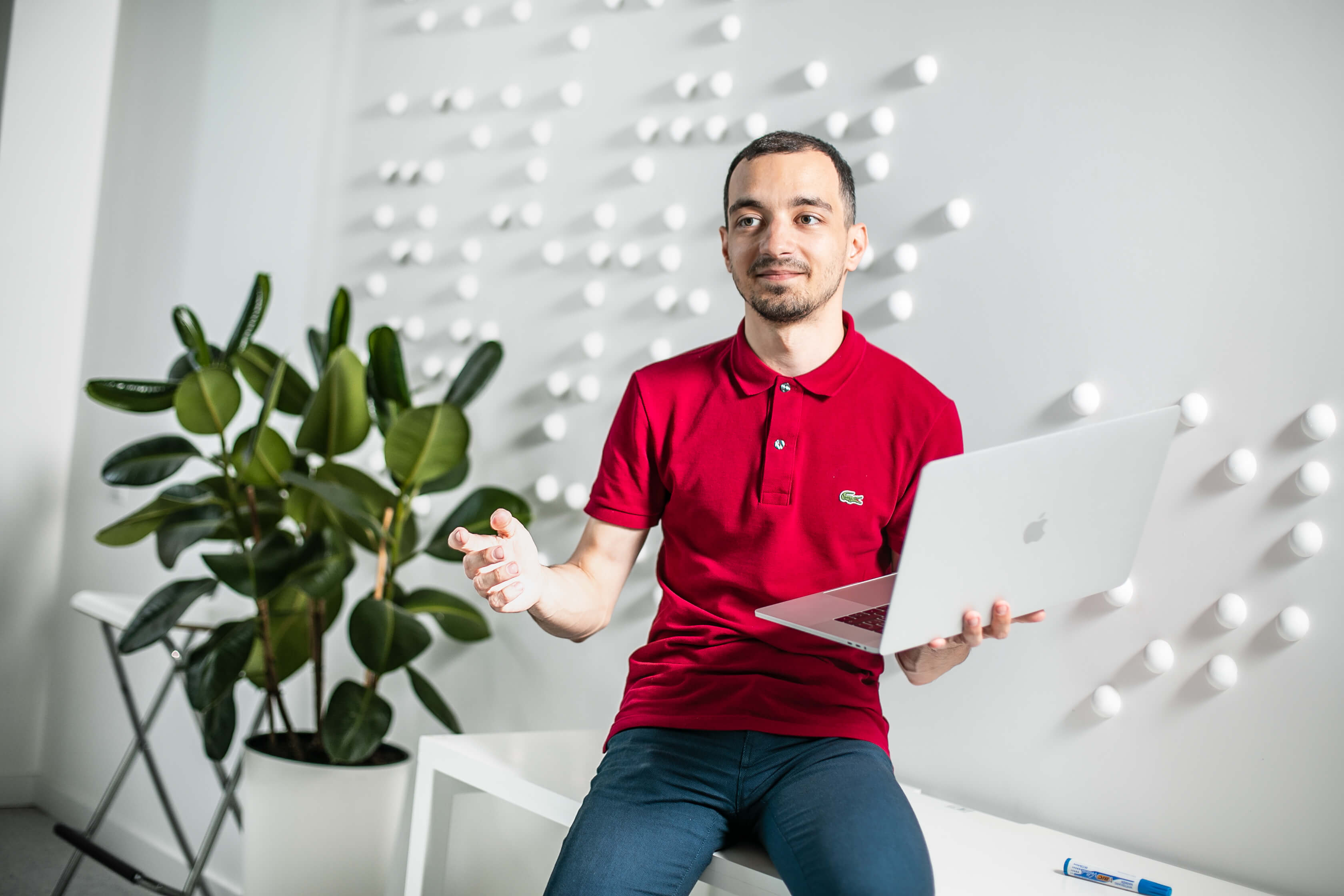 дизайнер Слава Шестопалов у червоній футболці сидить на столі з ноутбуком у руках та демонструє щось на його екрані, позаду нього — біла стіна та зелена рослина у горщику